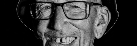 A black and white portrait of an older white man smiling wearing glasses and a Guinness bucket hat.