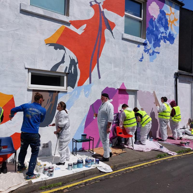A row of people in white overalls and yellow hi-vis vests painting a brightly coloured mural.